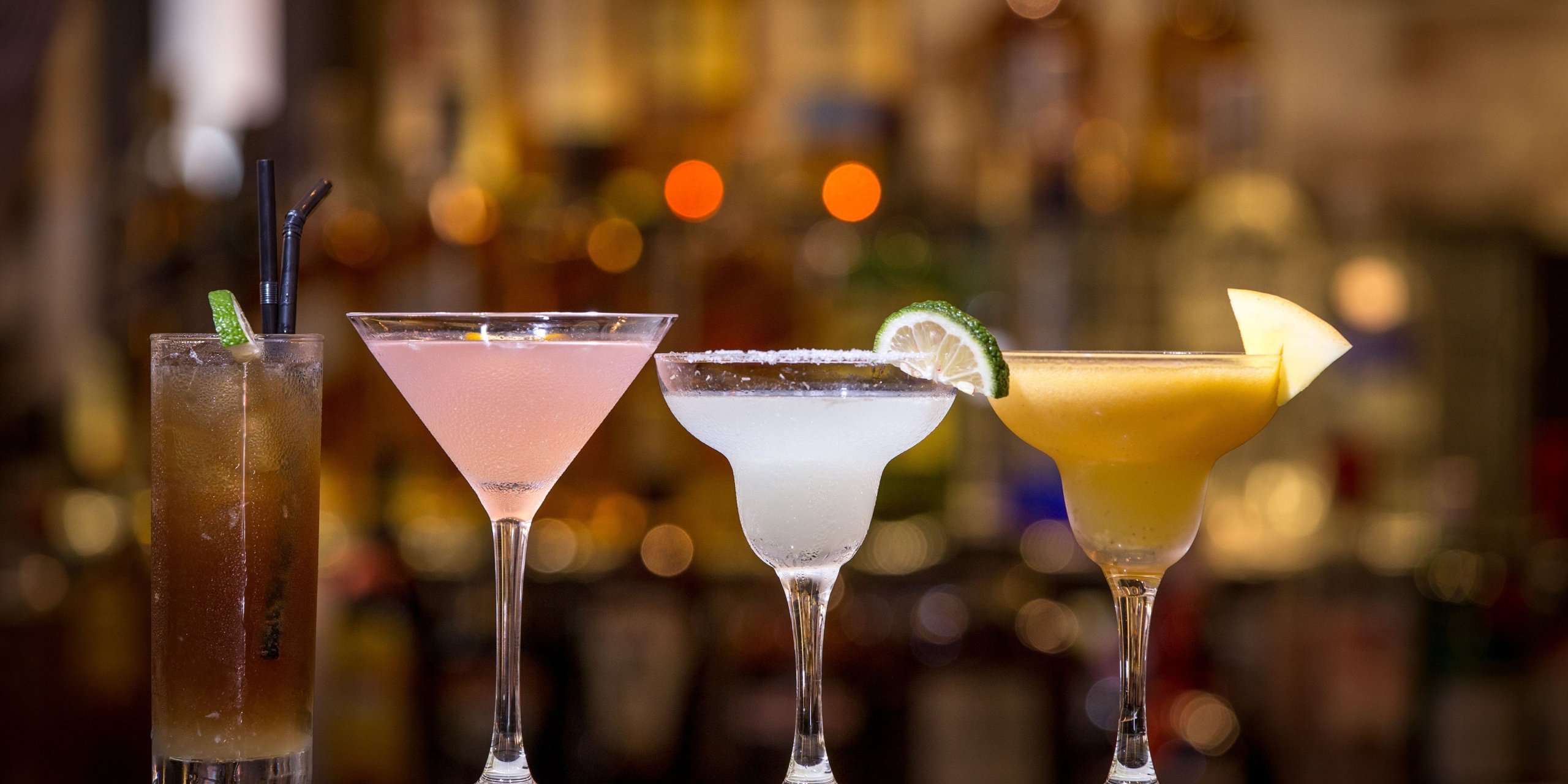 Four cocktails in a row on a bar counter, warm bar lights blurred behind them